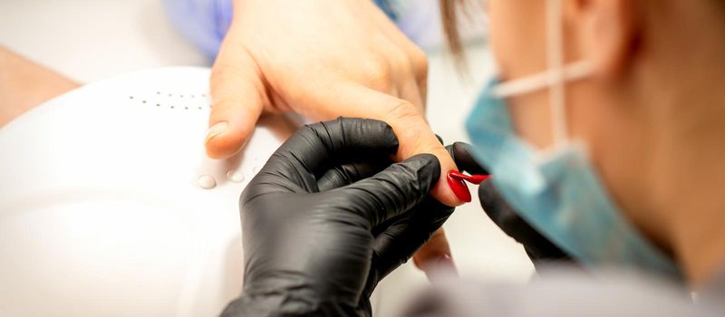 Manicure varnish painting. Close-up of a manicure master wearing rubber black gloves applying red varnish on a female fingernail in the beauty salon