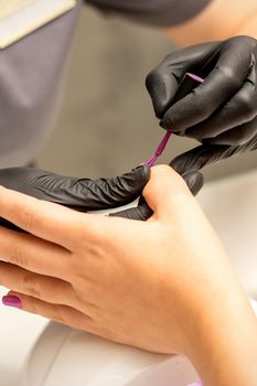 Professional manicure. A manicurist is painting the female nails of a client with purple nail polish in a beauty salon, close up. Beauty industry concept