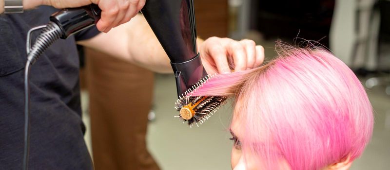 Drying short pink hair of young caucasian woman with a black hairdryer and black round brush by hands of a male hairdresser in a hair salon, close up