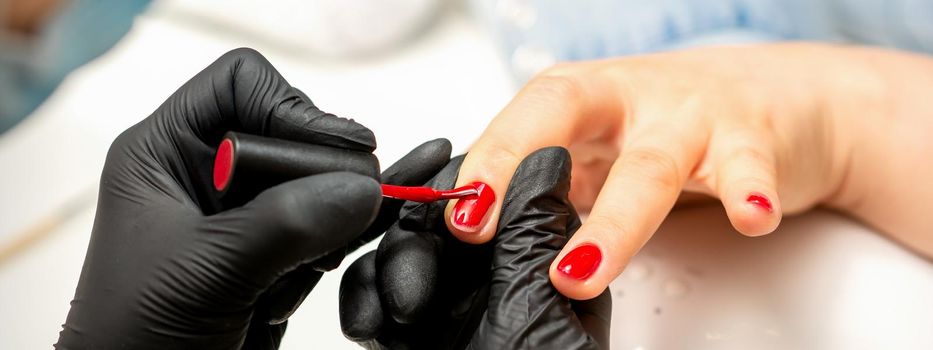 Manicure varnish painting. Close-up of a manicure master wearing rubber black gloves applying red varnish on a female fingernail in the beauty salon