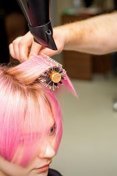 Drying short pink hair of young caucasian woman with a black hairdryer and black round brush by hands of a male hairdresser in a hair salon, close up