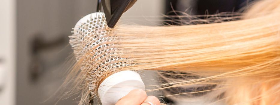 Drying straight blond hair with black hairdryer and white round brush in hairdresser salon, close up