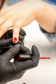 Manicure varnish painting. Close-up of a manicure master wearing rubber black gloves applying red varnish on a female fingernail in the beauty salon