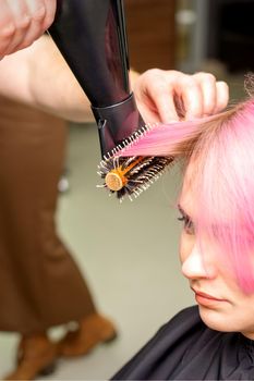 Drying short pink hair of young caucasian woman with a black hairdryer and black round brush by hands of a male hairdresser in a hair salon, close up