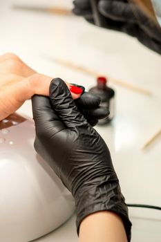 Manicure varnish painting. Close-up of a manicure master wearing rubber black gloves applying red varnish on a female fingernail in the beauty salon