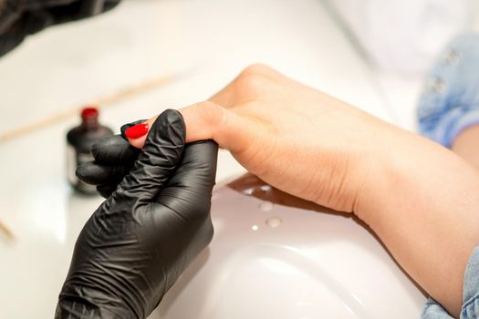 Manicure varnish painting. Close-up of a manicure master wearing rubber black gloves applying red varnish on a female fingernail in the beauty salon