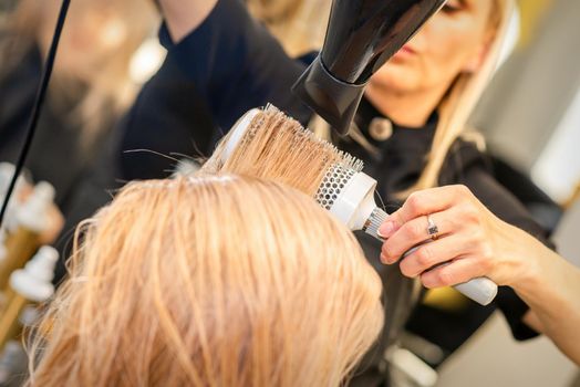 Drying straight blond hair with black hairdryer and white round brush in hairdresser salon, close up