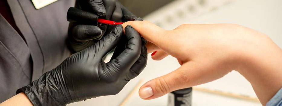 Manicure varnish painting. Close-up of a manicure master wearing rubber black gloves applying red varnish on a female fingernail in the beauty salon