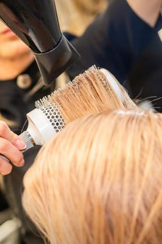 Drying straight blond hair with black hairdryer and white round brush in hairdresser salon, close up