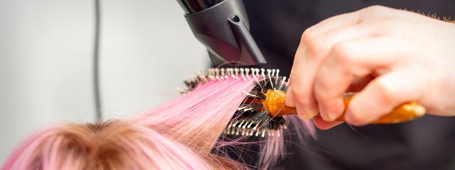 Drying short pink hair of young caucasian woman with a black hairdryer and black round brush by hands of a male hairdresser in a hair salon, close up