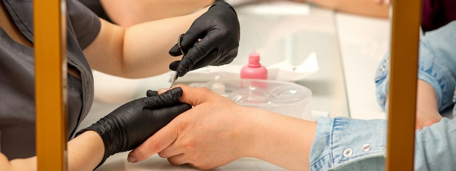 Manicure master removes cuticles from female nails with scissors wearing protective gloves in manicure salon