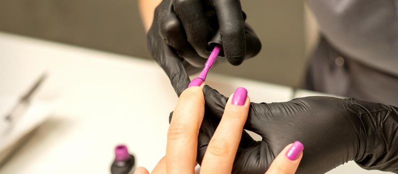 Professional manicure. A manicurist is painting the female nails of a client with purple nail polish in a beauty salon, close up. Beauty industry concept