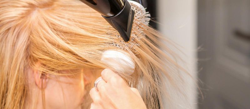 Drying straight blond hair with black hairdryer and white round brush in hairdresser salon, close up