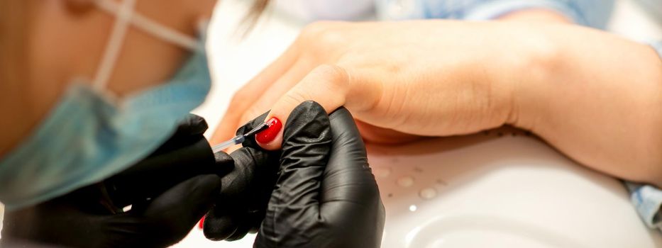 Professional manicure. A manicurist is painting the female nails of a client with red nail polish in a beauty salon, close up. Beauty industry concept