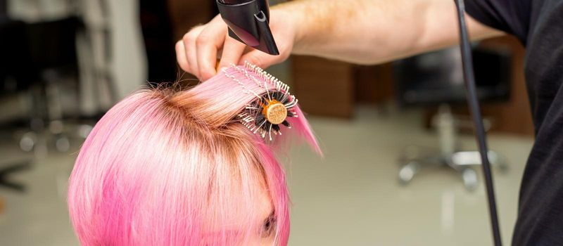 Drying short pink hair of young caucasian woman with a black hairdryer and black round brush by hands of a male hairdresser in a hair salon, close up