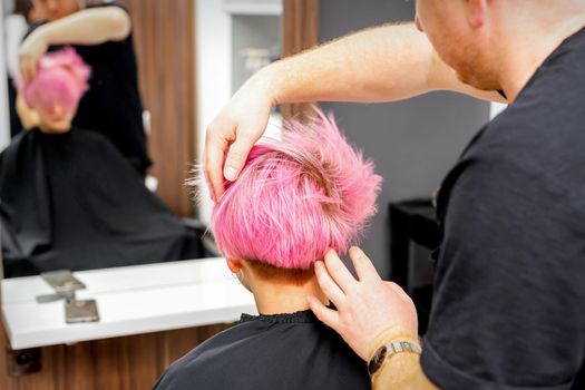 Hairdresser with hands is checking out and fixing the short pink hairstyle of the young white woman in a hair salon