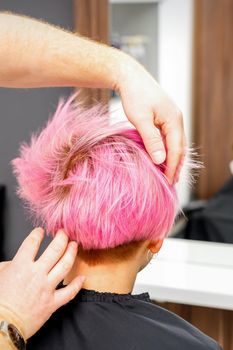 Hairdresser with hands is checking out and fixing the short pink hairstyle of the young white woman in a hair salon