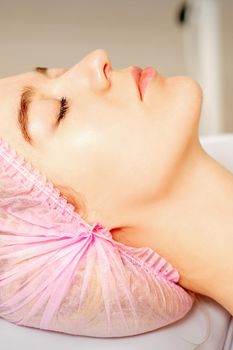 Side view of a young caucasian woman in a medical cap lying with closed eyes waiting for the cosmetic procedure in a beauty salon