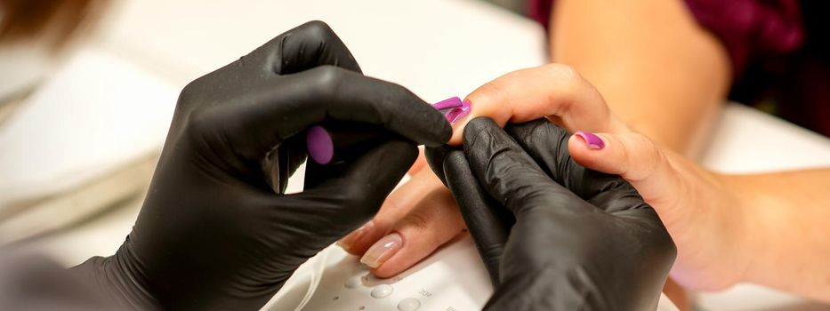 Professional manicure. A manicurist is painting the female nails of a client with purple nail polish in a beauty salon, close up. Beauty industry concept