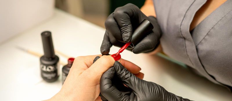 Professional manicure. A manicurist is painting the female nails of a client with red nail polish in a beauty salon, close up. Beauty industry concept