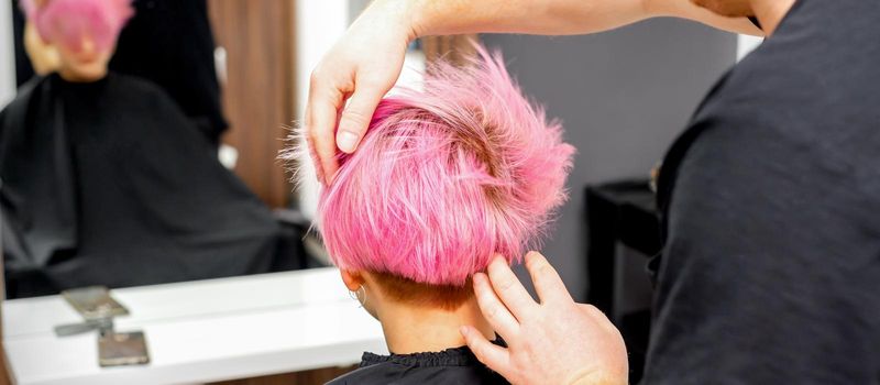 Hairdresser with hands is checking out and fixing the short pink hairstyle of the young white woman in a hair salon