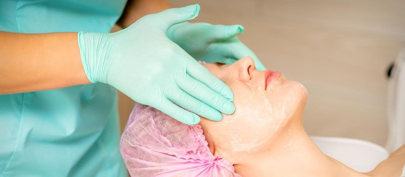 Cosmetologist with gloved hands applies a moisturizing mask with peeling cream on the female face. Facial cosmetology treatment. Procedures for facial care