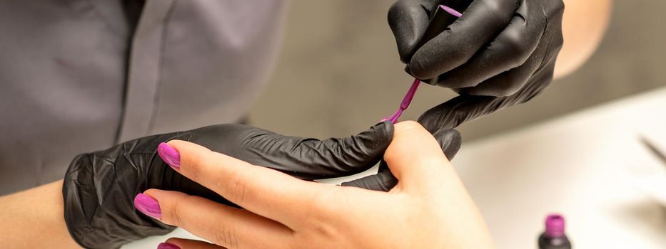 Professional manicure. A manicurist is painting the female nails of a client with purple nail polish in a beauty salon, close up. Beauty industry concept