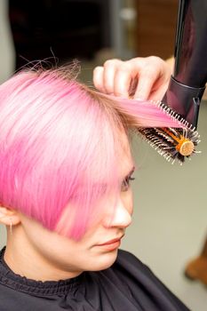 Drying short pink hair of young caucasian woman with a black hairdryer and black round brush by hands of a male hairdresser in a hair salon, close up