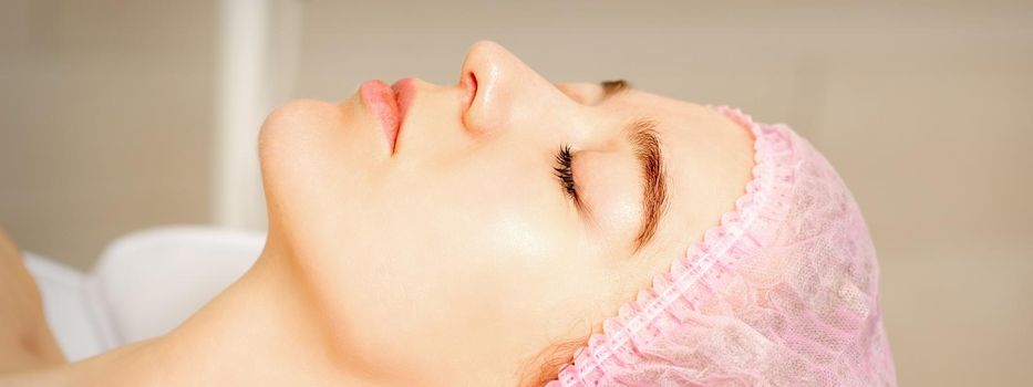 Side view of a young caucasian woman in a medical cap lying with closed eyes waiting for the cosmetic procedure in a beauty salon