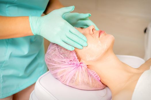 Cosmetologist with gloved hands applies a moisturizing mask with peeling cream on the female face. Facial cosmetology treatment. Procedures for facial care