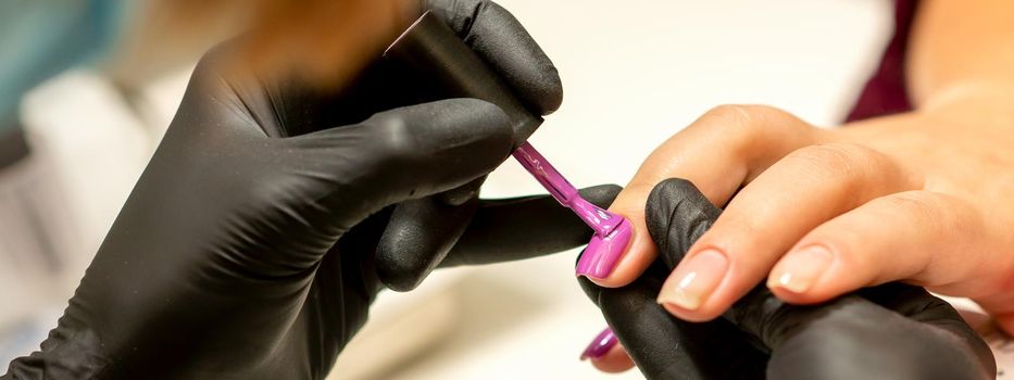 Professional manicure. A manicurist is painting the female nails of a client with purple nail polish in a beauty salon, close up. Beauty industry concept