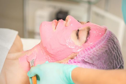 Hand of cosmetologist applying the pink alginic mask with the brush to the face of the young woman in a beauty salon