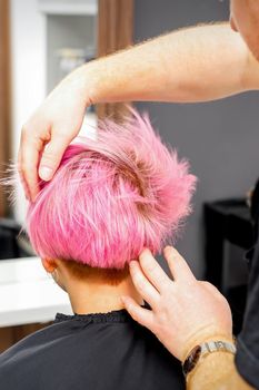 Hairdresser with hands is checking out and fixing the short pink hairstyle of the young white woman in a hair salon