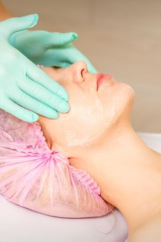 Cosmetologist with gloved hands applies a moisturizing mask with peeling cream on the female face. Facial cosmetology treatment. Procedures for facial care