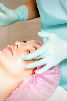 Cosmetologist with gloved hands applies a moisturizing mask with peeling cream on the female face. Facial cosmetology treatment. Procedures for facial care