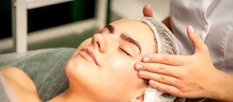 Head massage. Beautiful caucasian young white woman receiving a head and forehead massage with closed eyes in a spa salon