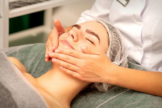 Beautiful caucasian young woman receiving a facial massage with closed eyes in spa salon, close up. Relaxing treatment concept