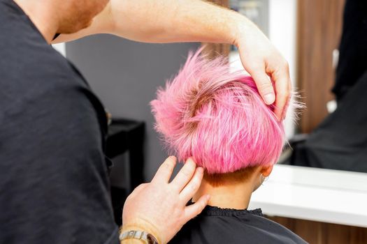 Hairdresser with hands is checking out and fixing the short pink hairstyle of the young white woman in a hair salon