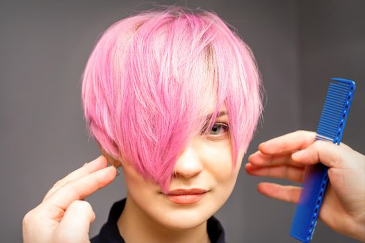 Hairdresser with hands and comb is checking out and fixing the short pink hairstyle of the young white woman in a hair salon