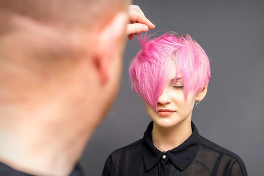 Hairdresser with hands is checking out and fixing the short pink hairstyle of the young white woman in a hair salon