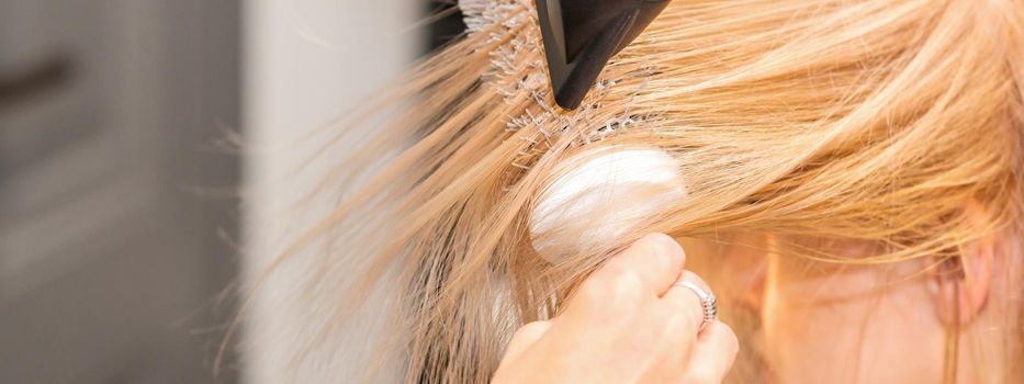 Drying straight blond hair with black hairdryer and white round brush in hairdresser salon, close up