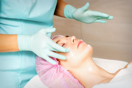 Cosmetologist with gloved hands applies a moisturizing mask with peeling cream on the female face. Facial cosmetology treatment. Procedures for facial care