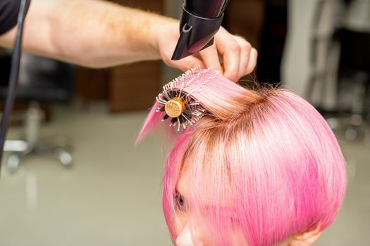 Drying short pink hair of young caucasian woman with a black hairdryer and black round brush by hands of a male hairdresser in a hair salon, close up
