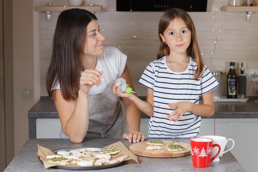 A little girl with her mother decorates Christmas cookies in the kitchen. Real life.