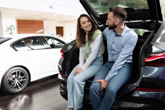 a young married couple in a car dealership chooses a car sits with a smile in the trunk of a crossover.