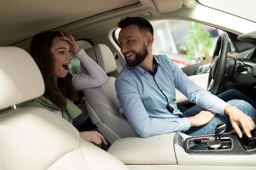 a man and a woman in a car dealership are sitting inside the car, being surprised by new technologies and promotions.