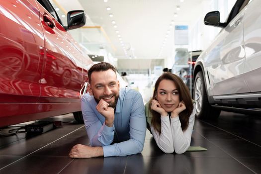 a cheerful married couple lies on the floor of a car dealership choosing a new car with a smile on their faces.