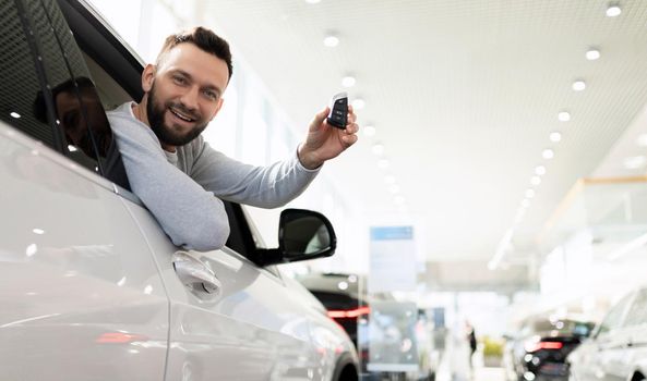 happy owner of a new car with a key in his hands in a dealership showroom looks out of the car.