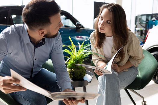 a young couple in a car dealership studying booklets with specifications and prices for new cars discussing buying a car.