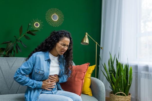Woman sick alone at home, Hispanic woman sitting on sofa in living room having severe stomach pain.